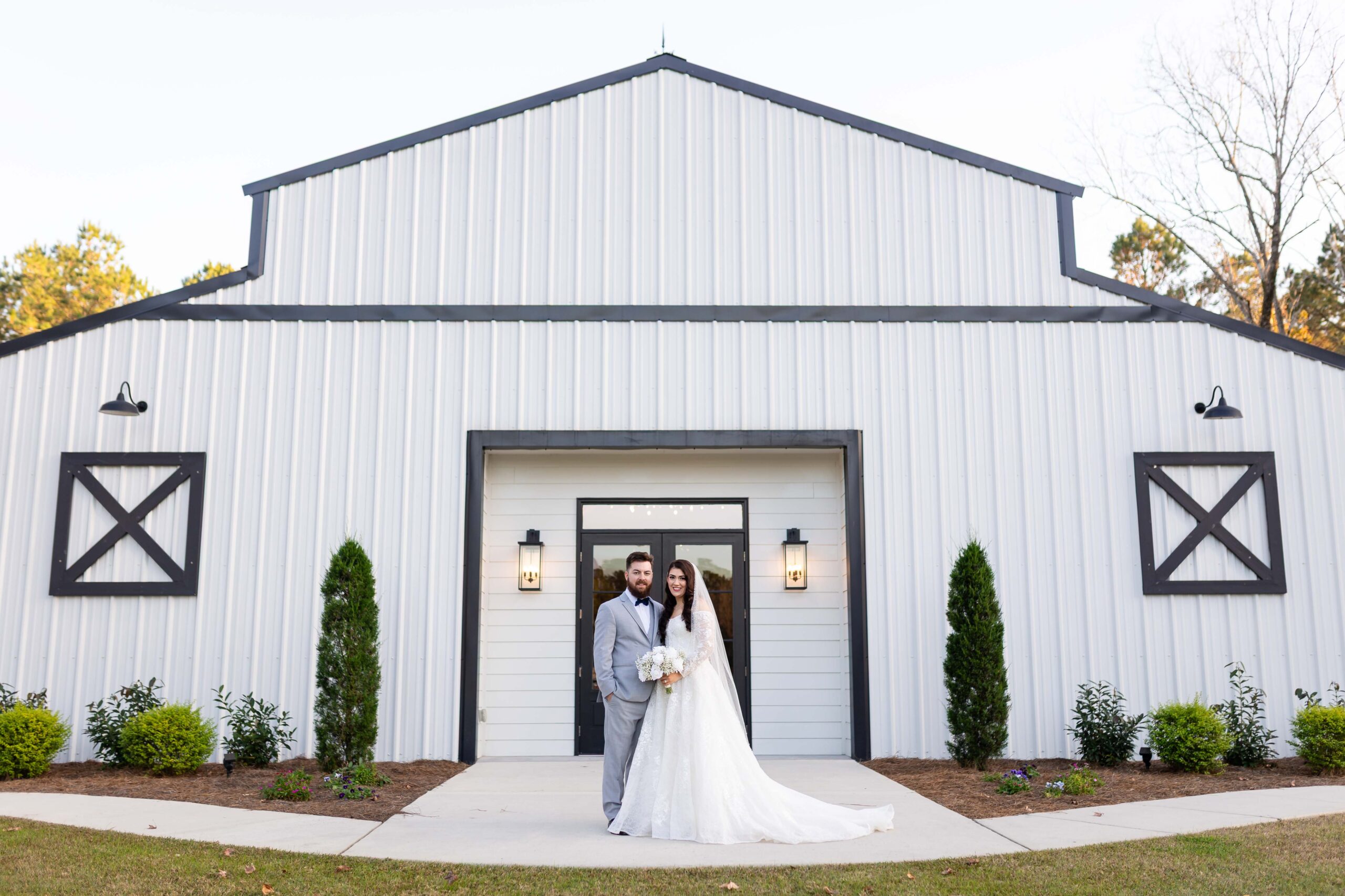 A bride and groom posing in front of The White Barn in Saucier, Mississippi. The couple is centered before a large white modern farmhouse-style barn with black trim and elegant outdoor lighting, photographed by Dear Magnolia Films