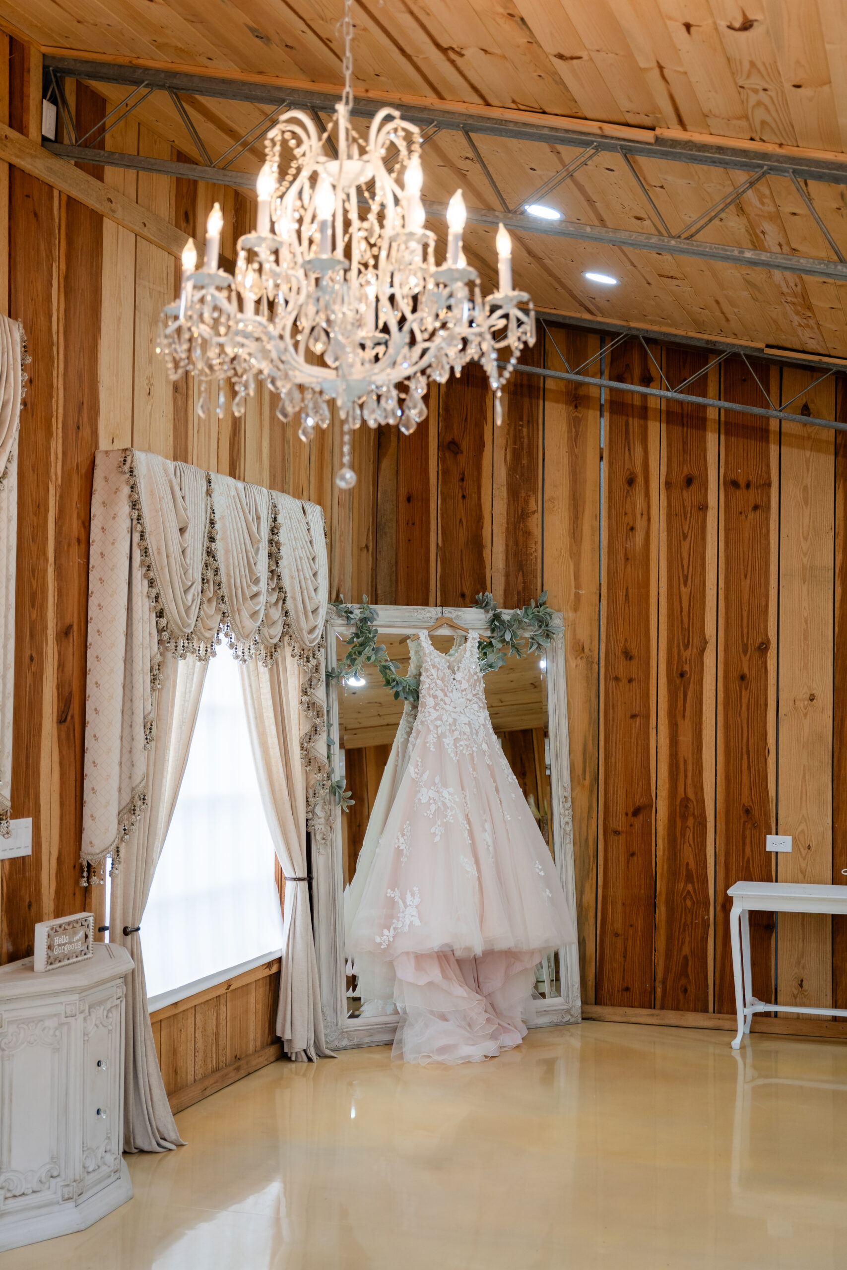 A detailed bridal photography shot of a wedding dress hanging on a floor-length mirror under a crystal chandelier at Rustic Grace Barn in Davenport Florida by Dear Magnolia Films.