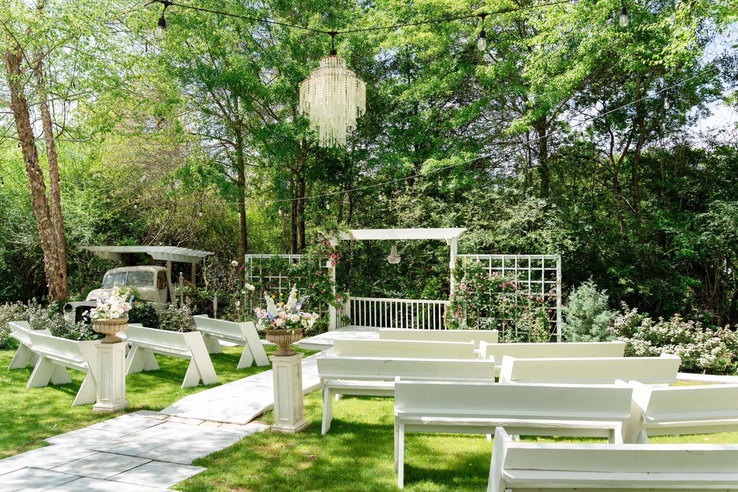 Outdoor wedding ceremony setup at La Petite Venue featuring white wooden benches, a matching white altar with pink climbing roses, and a crystal chandelier hanging from a string of bistro lights against a lush green forest backdrop.