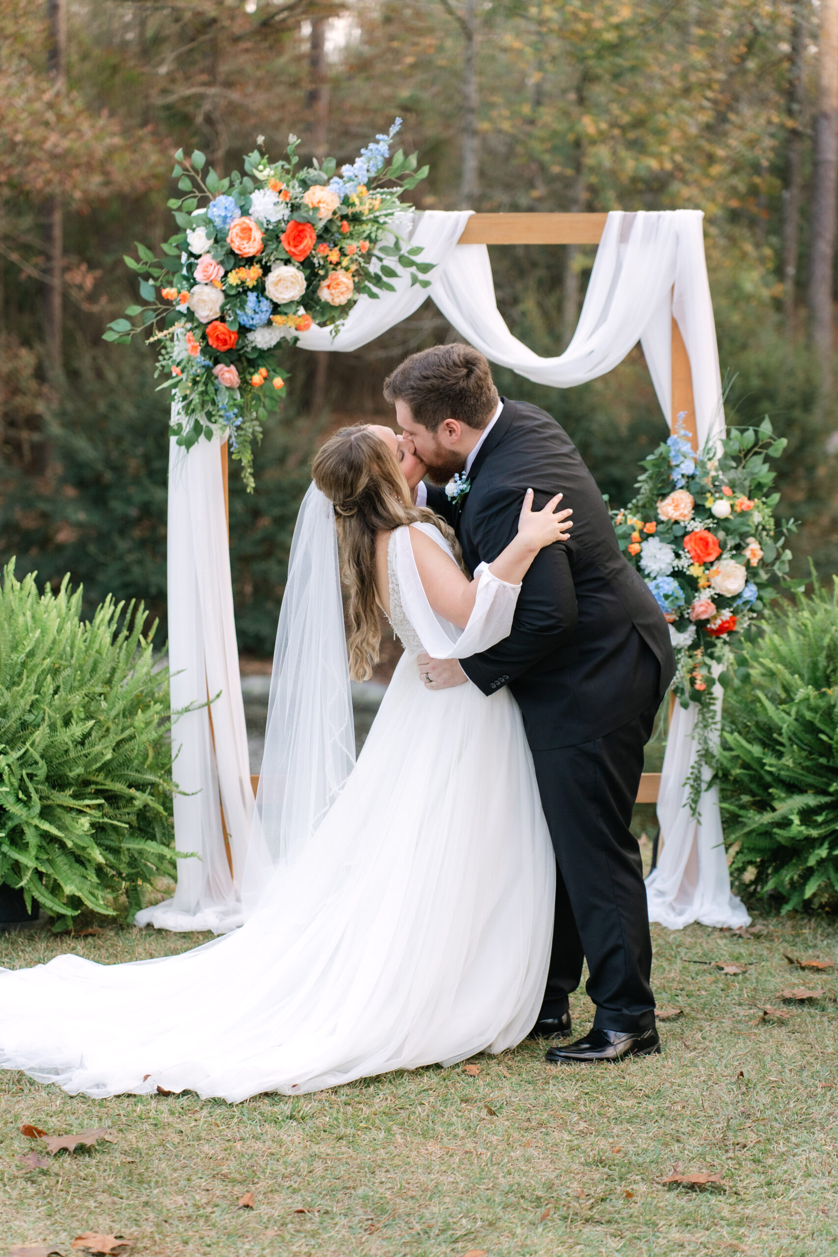 A newly married couple shares their first kiss at Dogwood Venue in Sumrall, MS. The outdoor ceremony features a wooden arbor draped in white fabric with vibrant orange and blue floral arrangements, captured by Dear Magnolia Films.