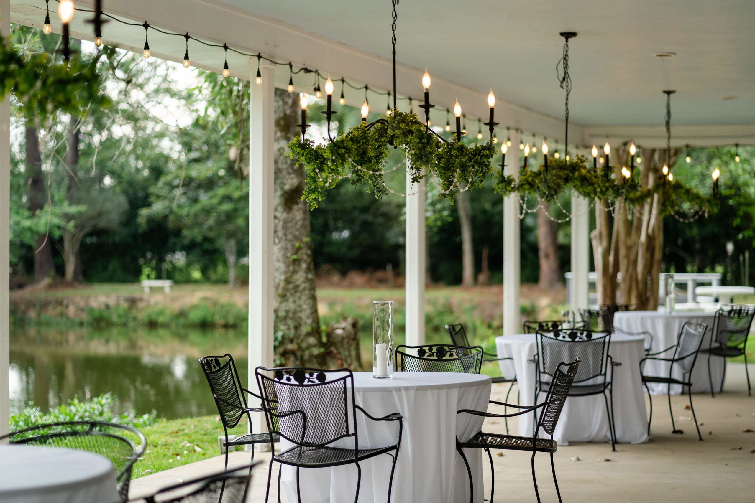 Outdoor reception patio at Lynhaven Events in Hammond LA featuring white-tablecloth tables, wrought-iron chairs, and greenery-wrapped chandeliers over a pond view by Dear Magnolia Films.