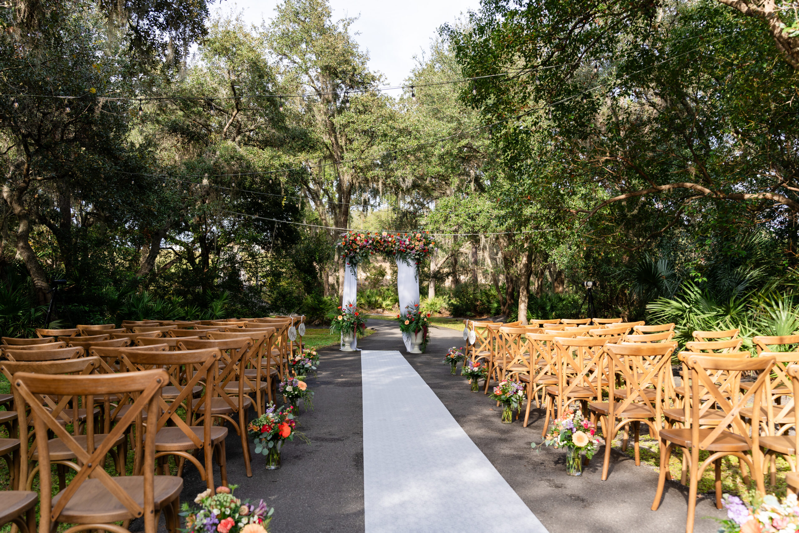 Outdoor wedding ceremony altar at Rustic Grace Barn in Davenport FL featuring a wooden cross, floral arch, and rustic log benches under moss-draped oak trees by Dear Magnolia Films.